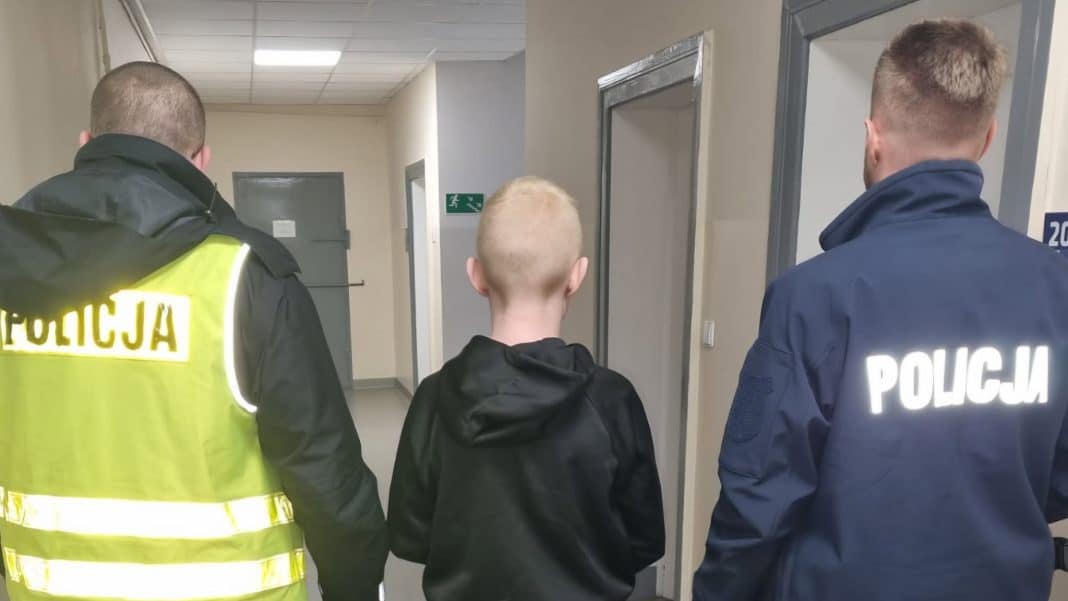 Policjanci prowadzą młodego chłopca w korytarzu szkoły, z tyłu widoczne drzwi i ściana.