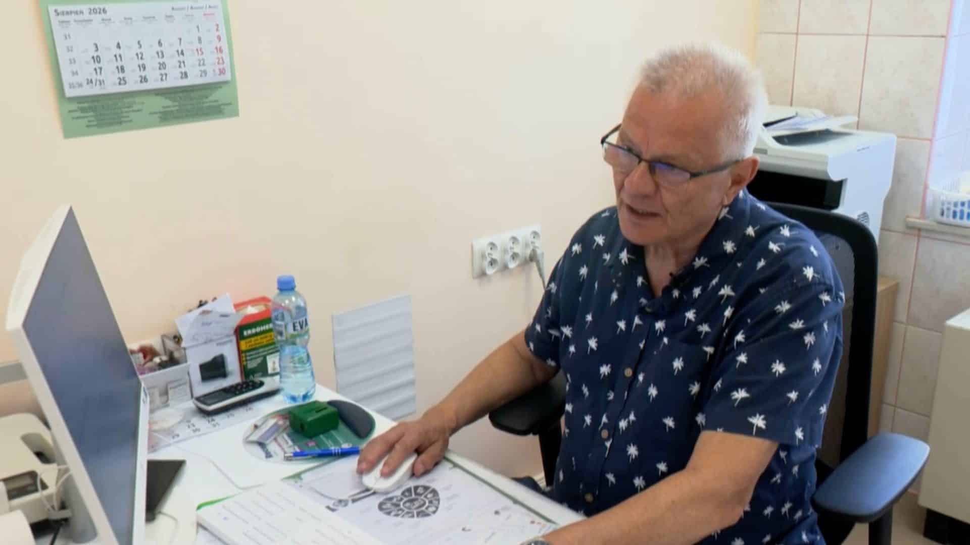 Mężczyzna siedzi przy biurku w gabinecie, patrzy na komputer, obok są lekarstwa i butelka wody.