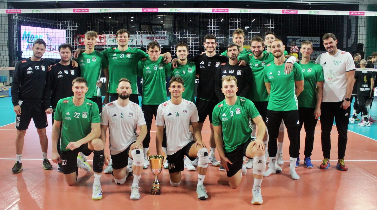 Grupa siatkarzy i trenerów AZS Olsztyn na boisku, z trofeum w centrum, podczas prezentacji drużyny.