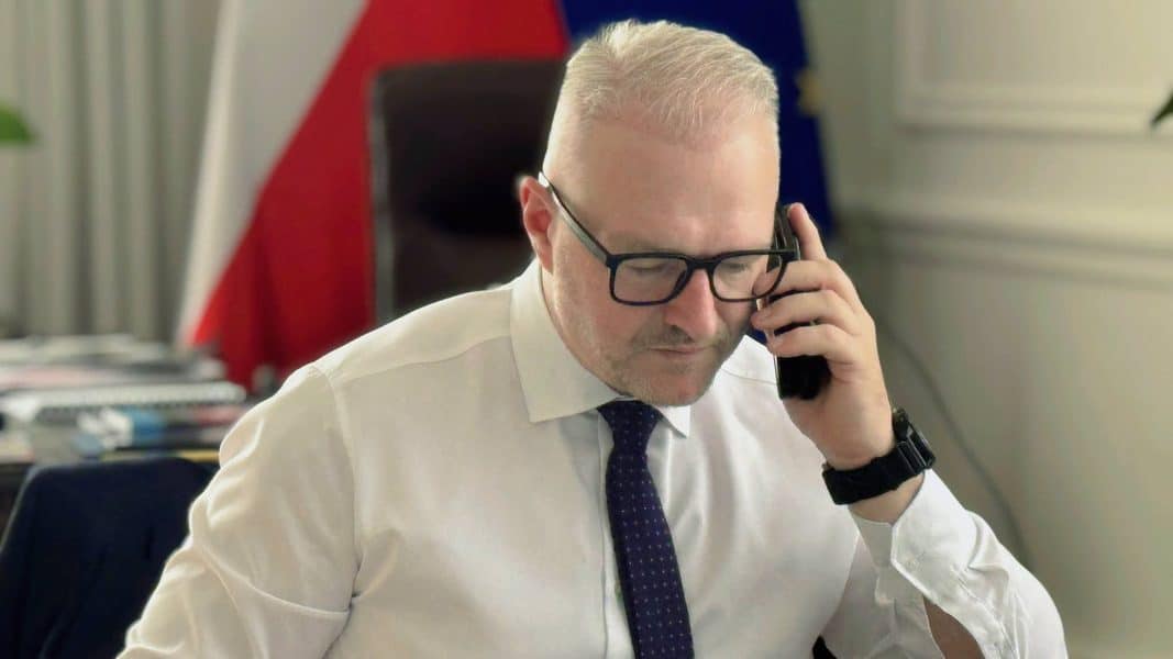 Mężczyzna w białej koszuli i okularach rozmawia przez telefon, siedząc przy biurku.