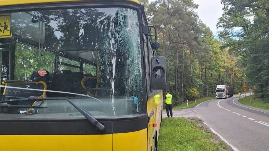 Uszkodzony autobus szkolny z widocznym pęknięciem na przedniej szybie, na tle lasu i policjantów.