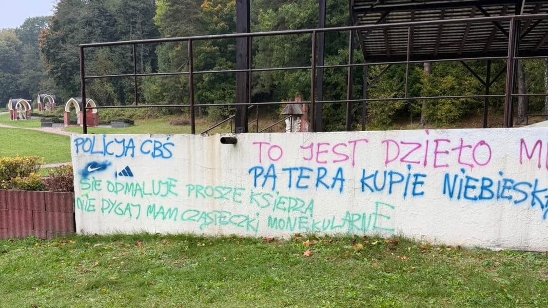 Mural na ścianie z napisem i graffiti, w tle teren zielony i konstrukcje na zewnątrz.