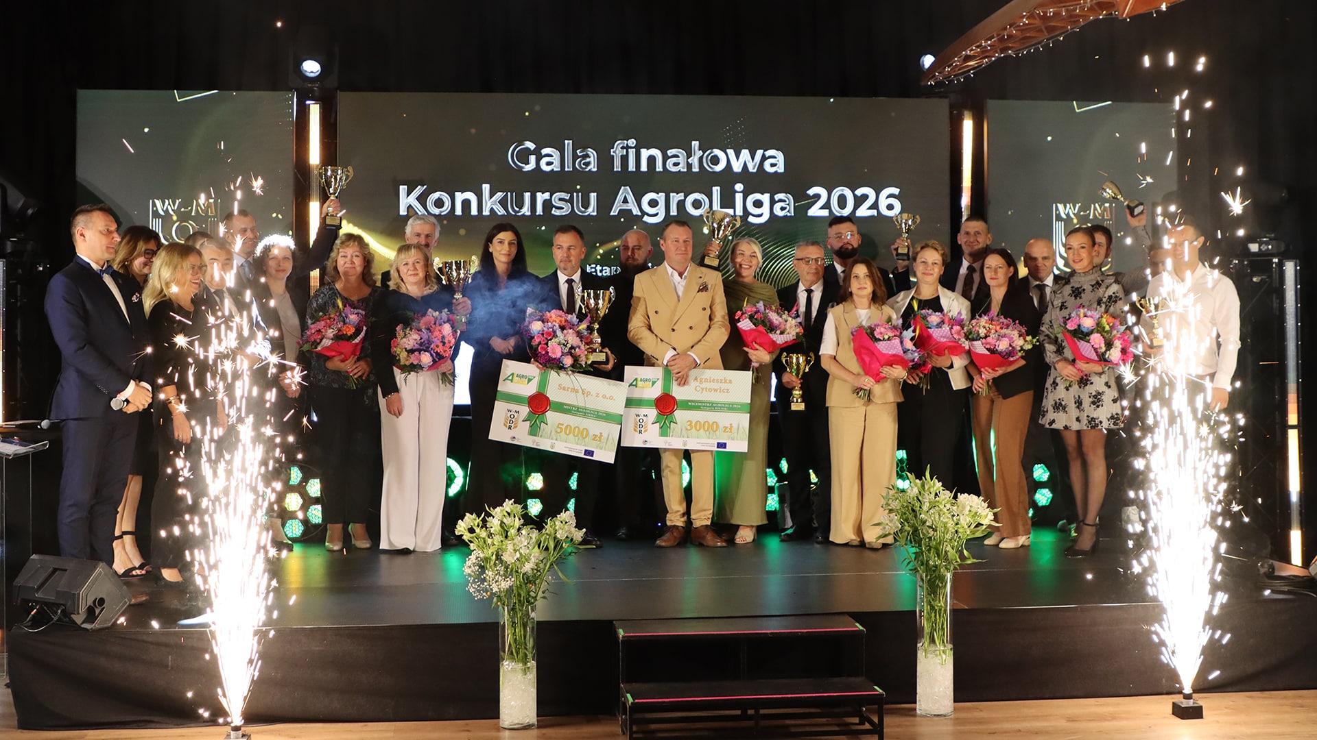 Grupa wyróżnionych rolników i firm na gali finałowej AgroLiga 2026, z trofeami i bukietami kwiatów.