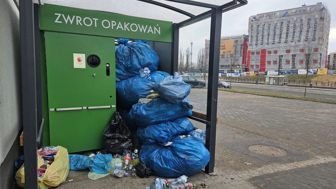 Stacja zwrotu opakowań z dużą ilością odpadów w workach i na ziemi, w tle budynek pod remont.