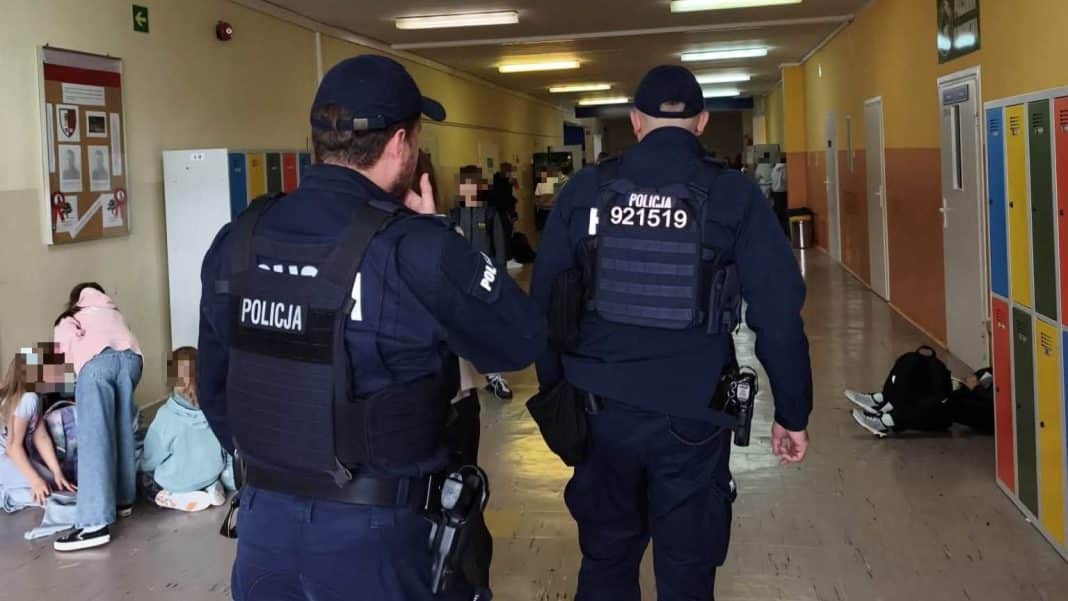 Policjanci patrolują korytarz szkolny, dzieci siedzą na podłodze przy szafkach.