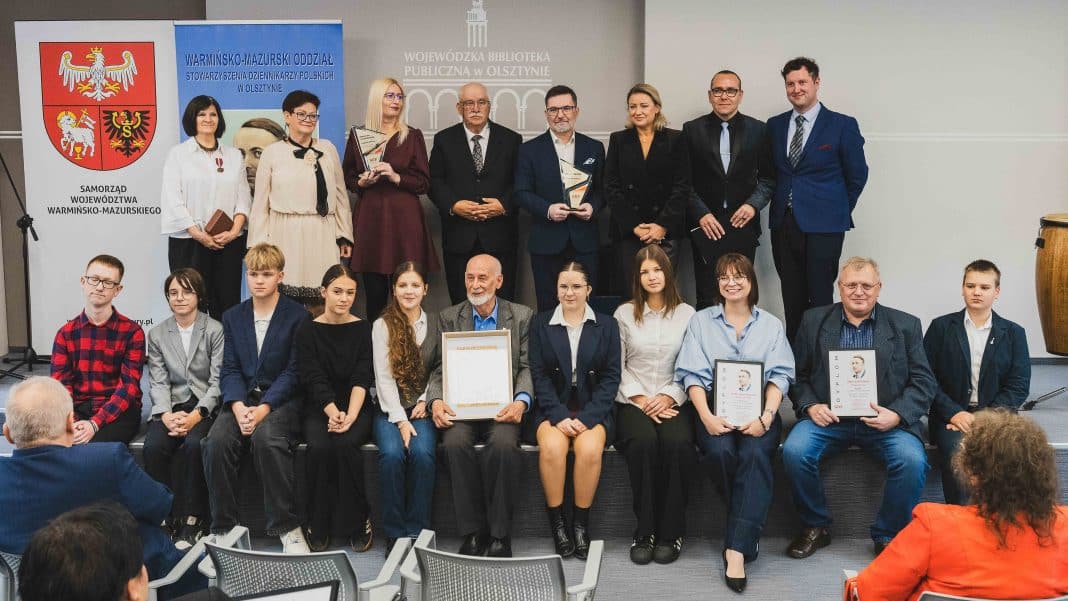 Grupa laureatów i organizatorów podczas gali w Olsztynie, na tle bannerów i logotypów wydarzenia.