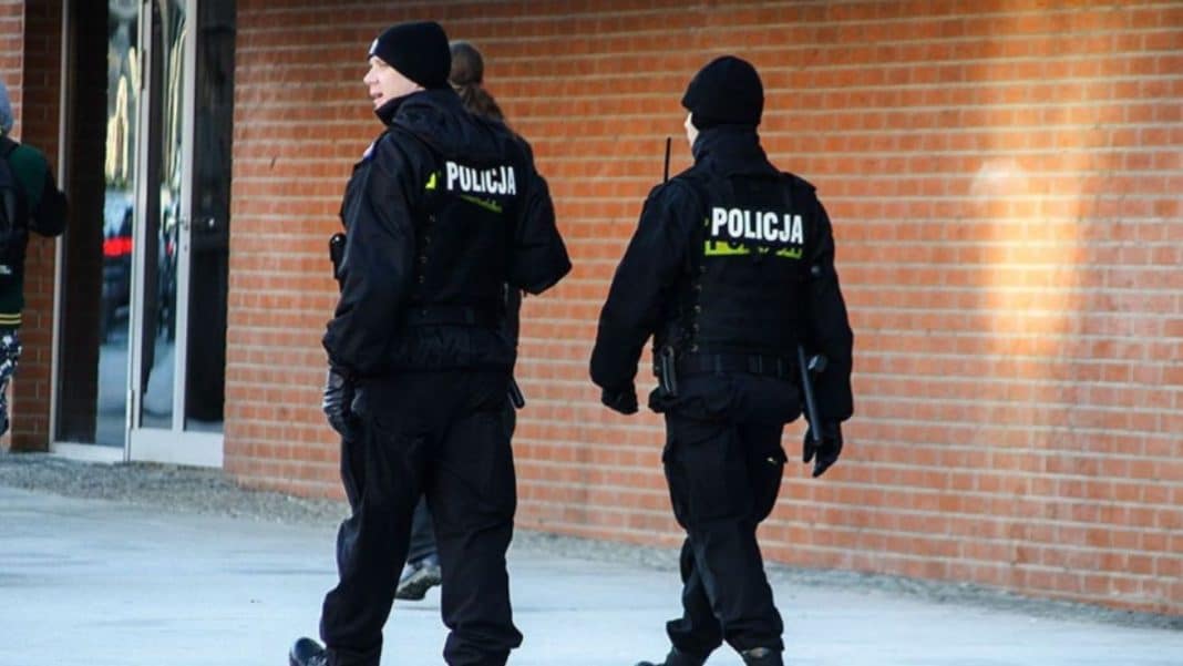 Dwoje policjantów w czarnych mundurach na tle ceglanej ściany, idących w kierunku wejścia do budynku.