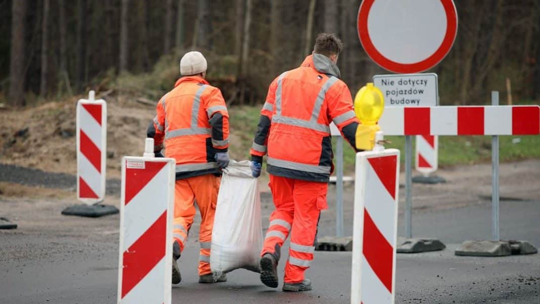 Pracownicy w odblaskowych kamizelkach pracują na zamkniętym skrzyżowaniu, otoczeni barierkami i znakami drogowymi.