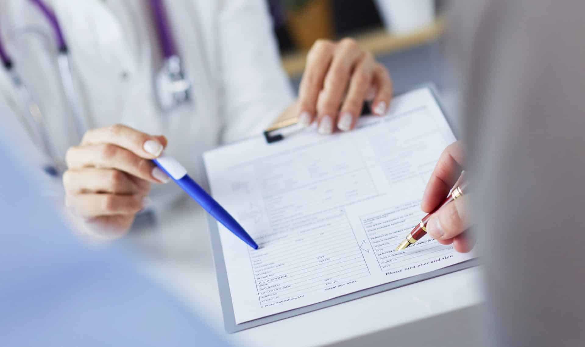 Osoba w białym kitlu wypełnia formularz w placówce medycznej, trzymając długopis i dokumenty.