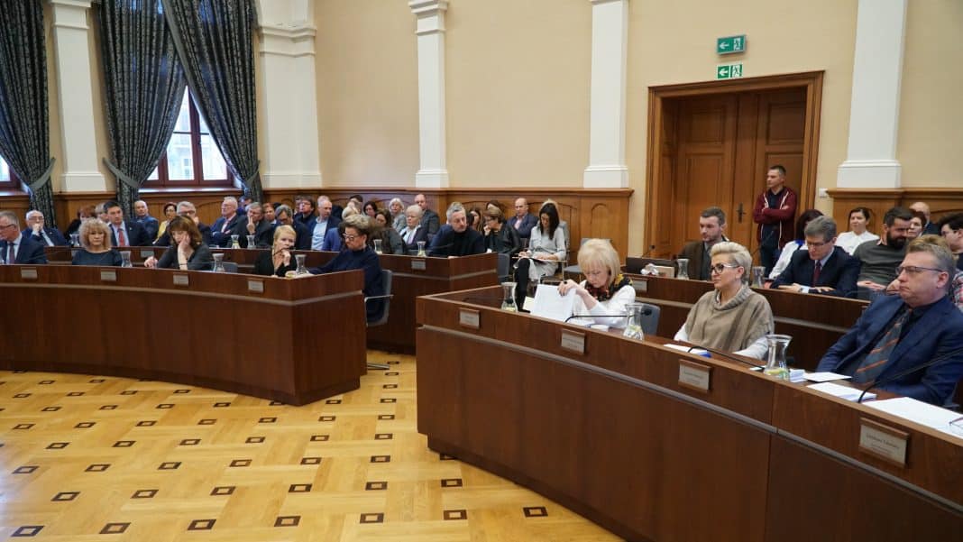 Radni siedzą przy długich, drewnianych stołach w dużej sali obrad, w tle okna z zasłonami i drzwi.