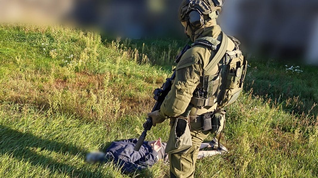 Żołnierz w kamuflażu na polu, trzymający broń, obok leży ciało, w tle zieleń i krzewy.