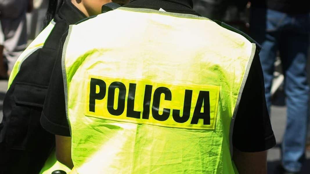 Policjant w kamizelce odblaskowej z napisem POLICJA na tle grupy ludzi.