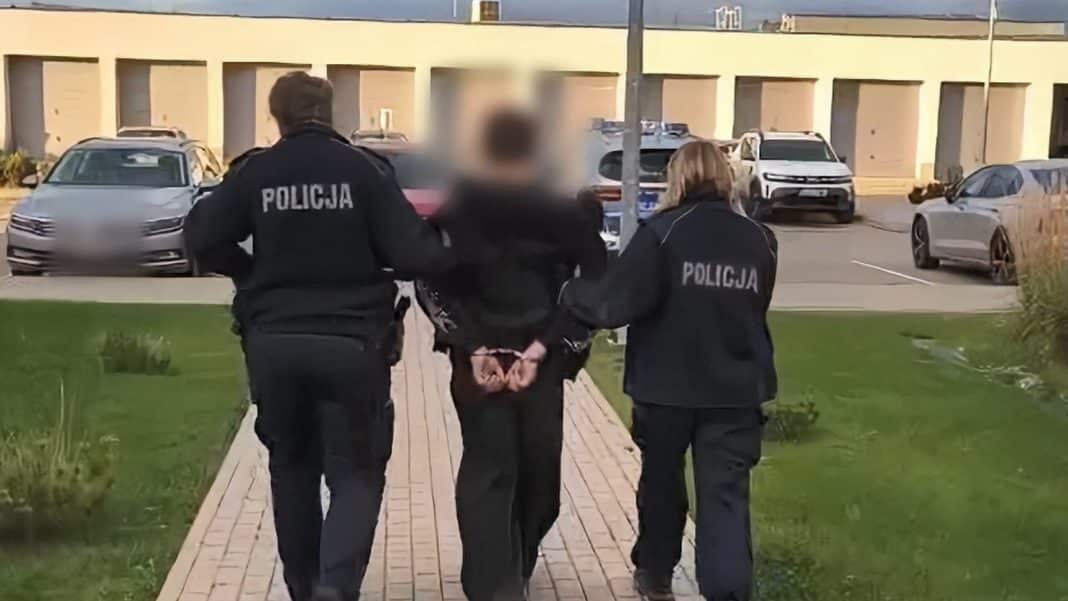 Policjanci prowadzą zatrzymanego chłopca na ulicy, w tle samochody i budynek.