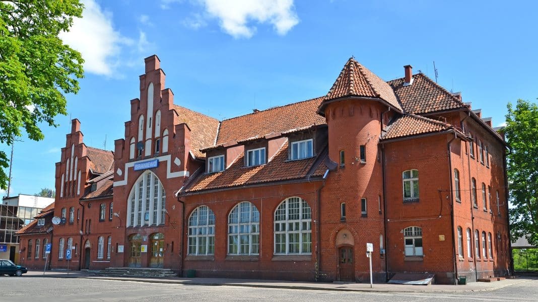 Zabytkowy, czerwony budynek dworca kolejowego z dużymi oknami i wieżyczkami, otoczony zielonymi drzewami, na tle nieba.