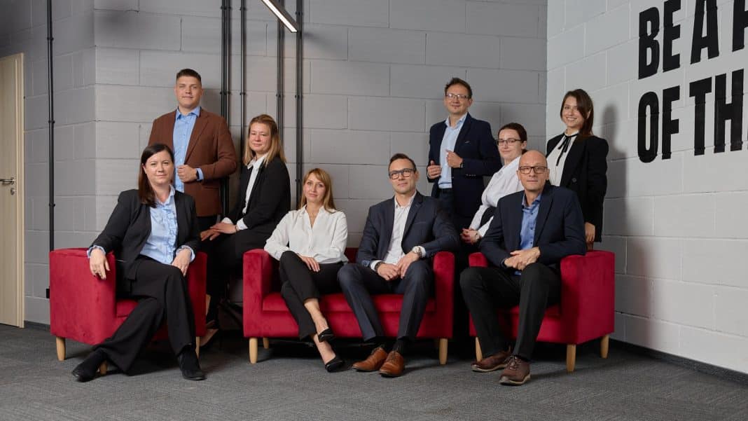 Grupa ludzi w formalnych ubraniach na tle szarej ściany, niektórzy siedzą na czerwonych fotelach, inni stoją.