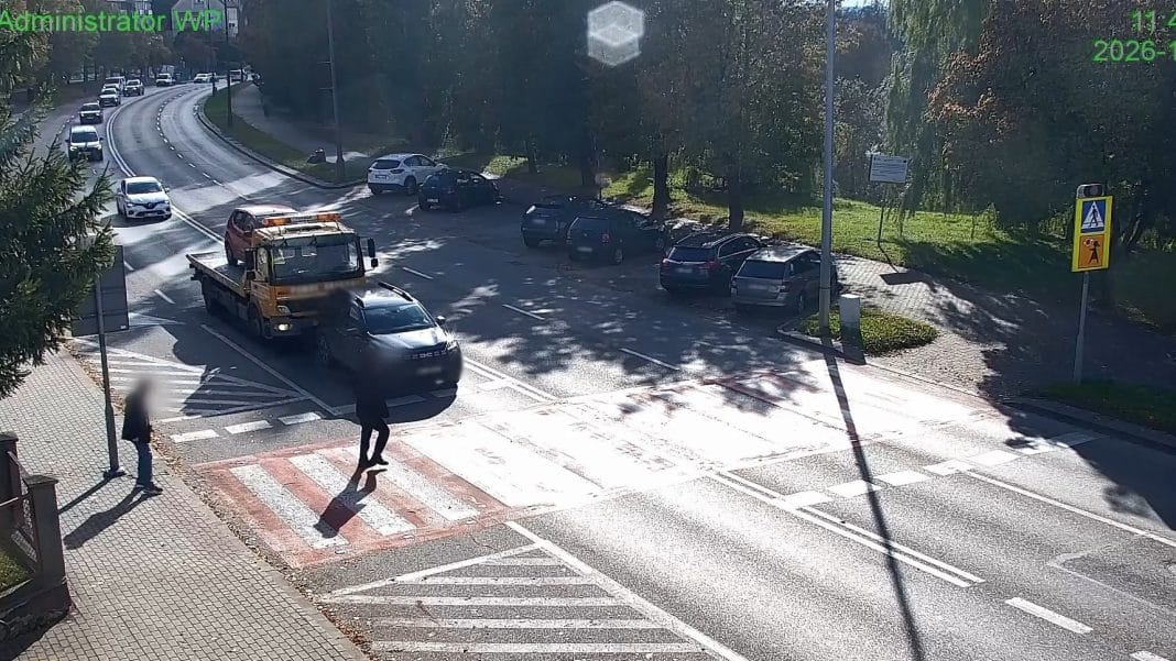 Zdjęcie z monitoringu pokazujące samochody na ulicy, pieszych na przejściu i cienie drzew.