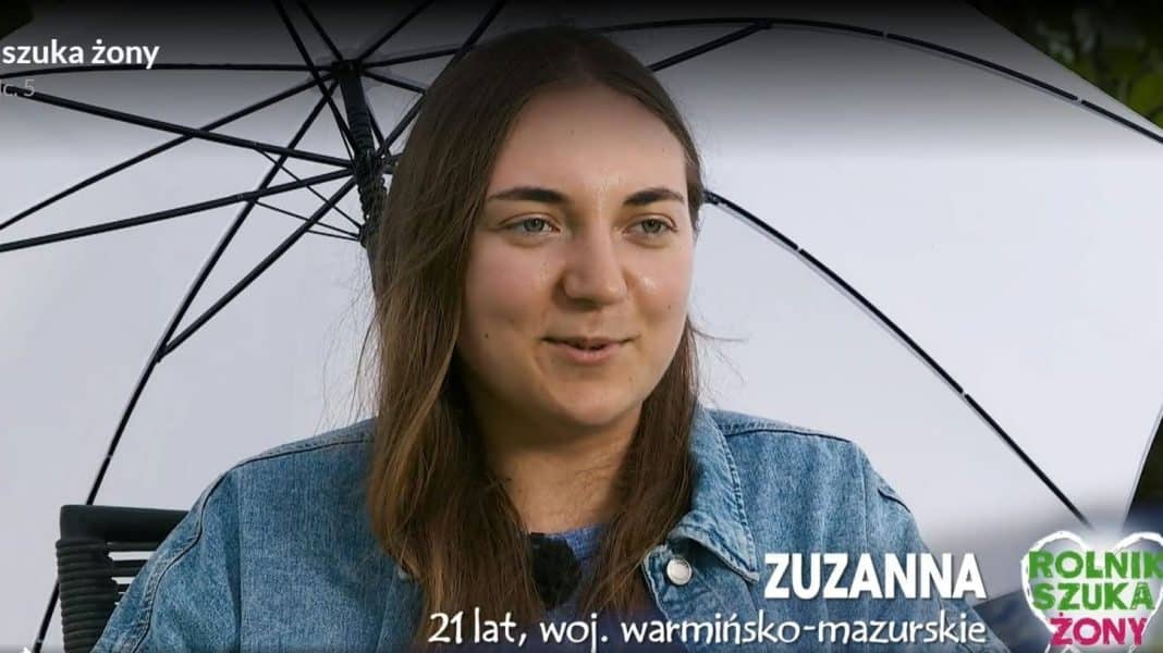 Młoda kobieta z długimi brązowymi włosami, trzymająca parasol, uśmiecha się podczas wywiadu na zewnątrz.