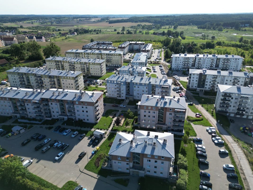Widok na osiedle bloków mieszkalnych z parkingami i zielonymi terenami wokół, w tle pola i krajobraz wiejski.