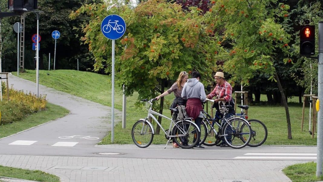 Trzy osoby z rowerami czekają na przejściu dla pieszych pod światłem czerwonym, w tle drzewa i ścieżki rowerowe.