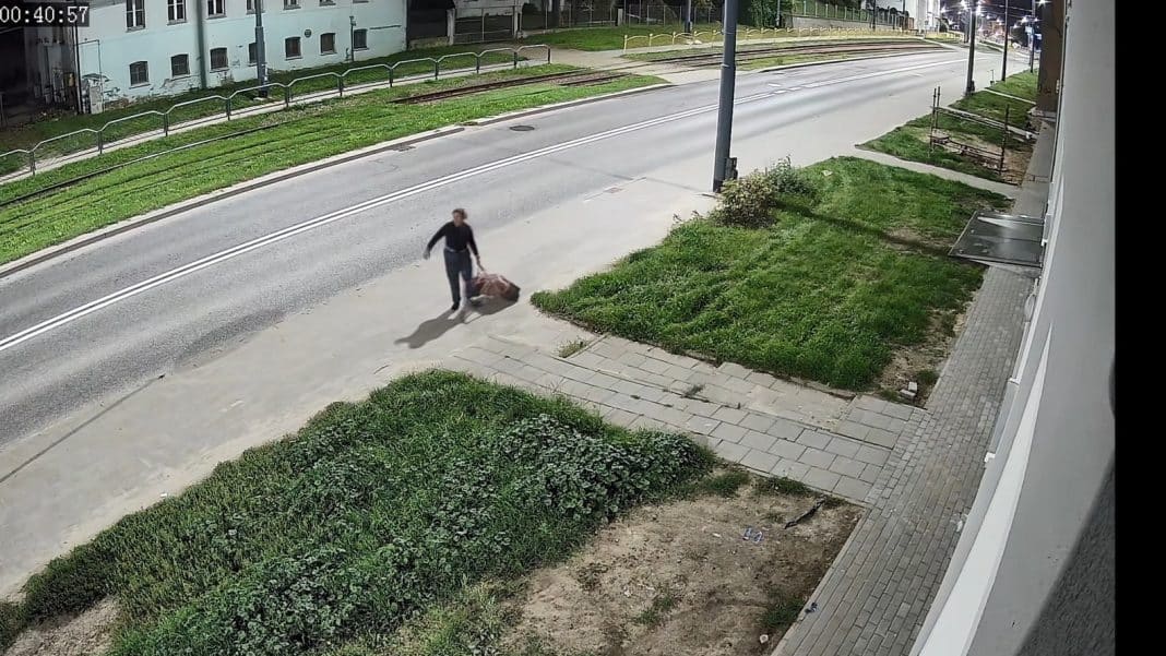 Mężczyzna z walizką na chodniku przy ulicy, w tle ulica i budynki, dzień.