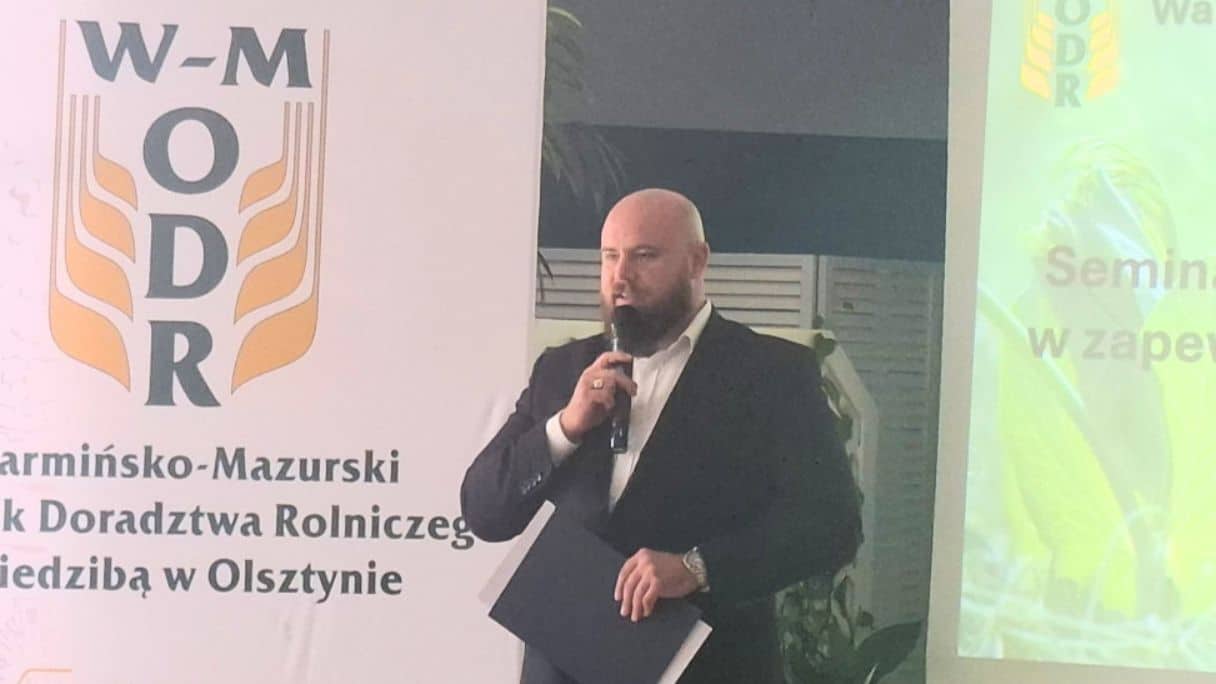 Mężczyzna w garniturze z mikrofonem podczas seminarium o roślinach bobowatych, w tle logo WMODR.