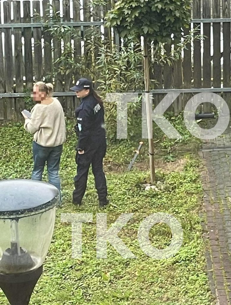 Policjantka i kobieta stoją na trawniku, obok wbita w ziemię maczeta, w tle drewniany płot i rośliny.