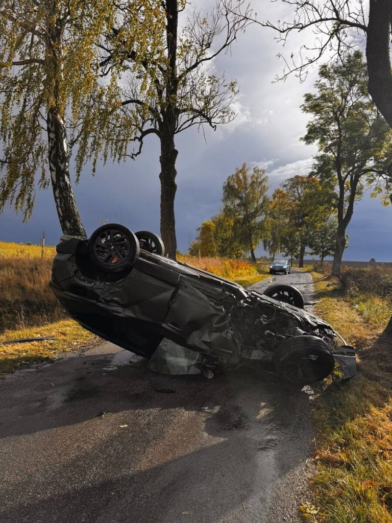 Rozbity samochód leży na boku przy drodze wśród drzew, w tle chmury i inne pojazdy.