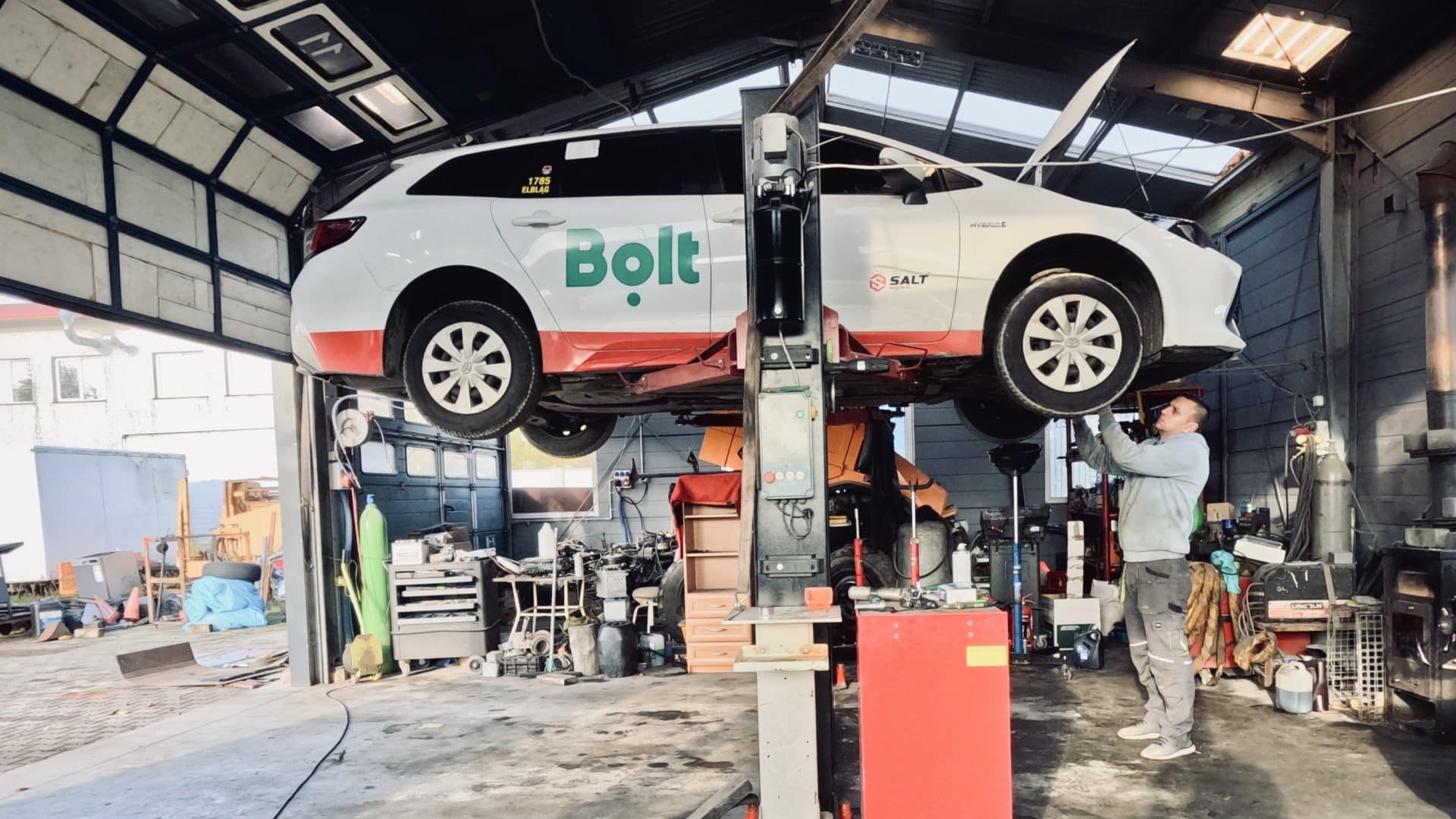 Wewnątrz warsztatu samochodowego, podniesiony samochód z logo Bolt, mechanik pracuje przy podwoziu.