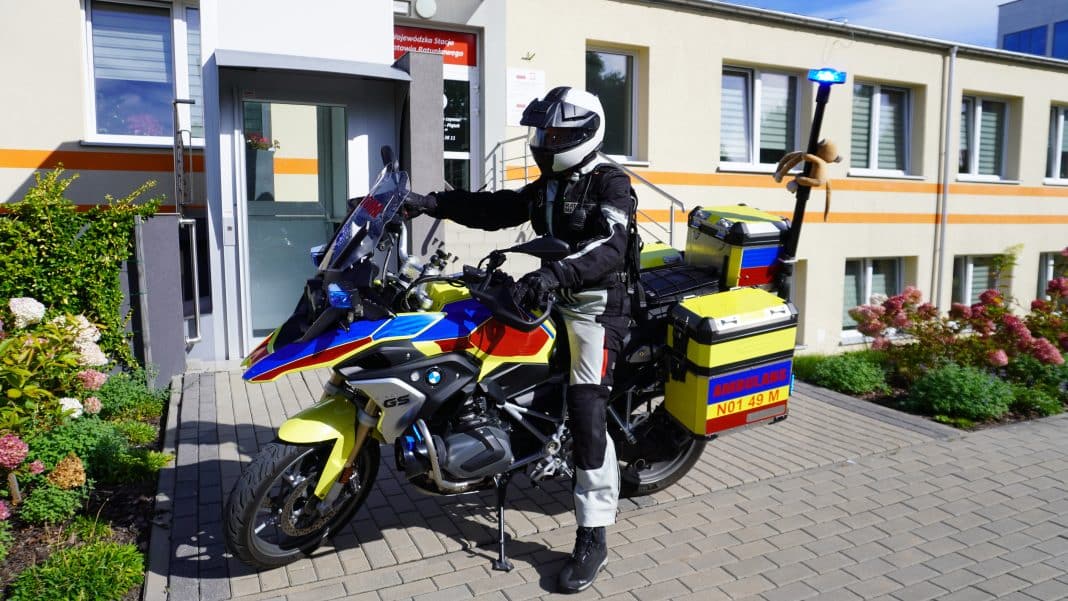 Ratownik na motocyklu ratunkowym stoi przed budynkiem, w pełnym wyposażeniu, z niebieskim światłem włączonym.