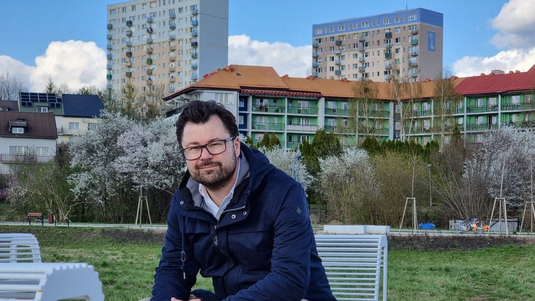 Mężczyzna siedzi na ławce w parku z drzewami w kwitnącym okresie i wysokimi budynkami w tle.