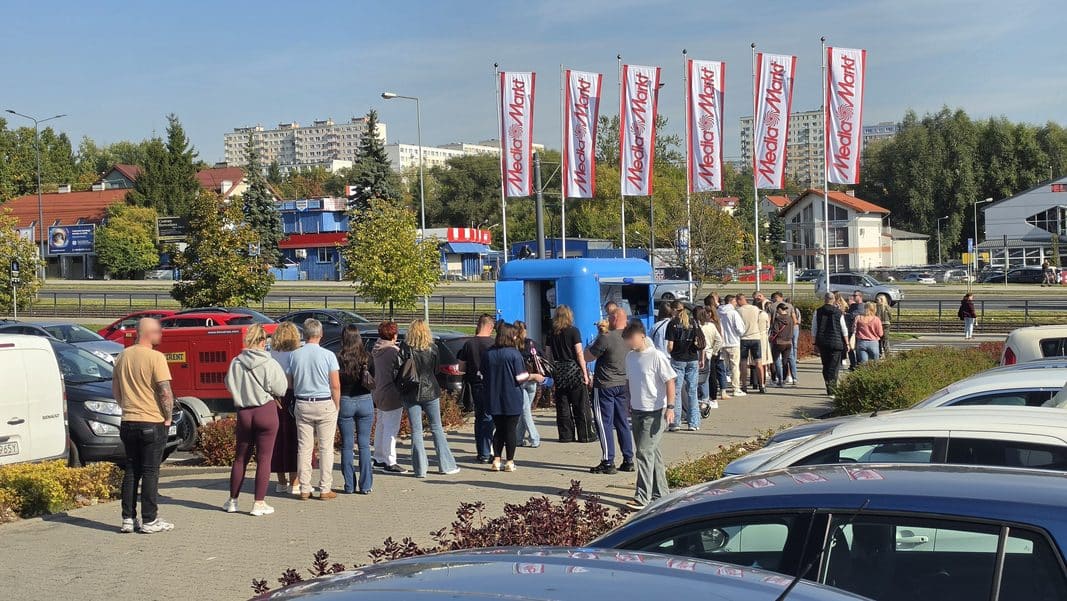 Długi kolejka ludzi czekających na wejście do lokalu w Olsztynie, w tle parking i budynki.