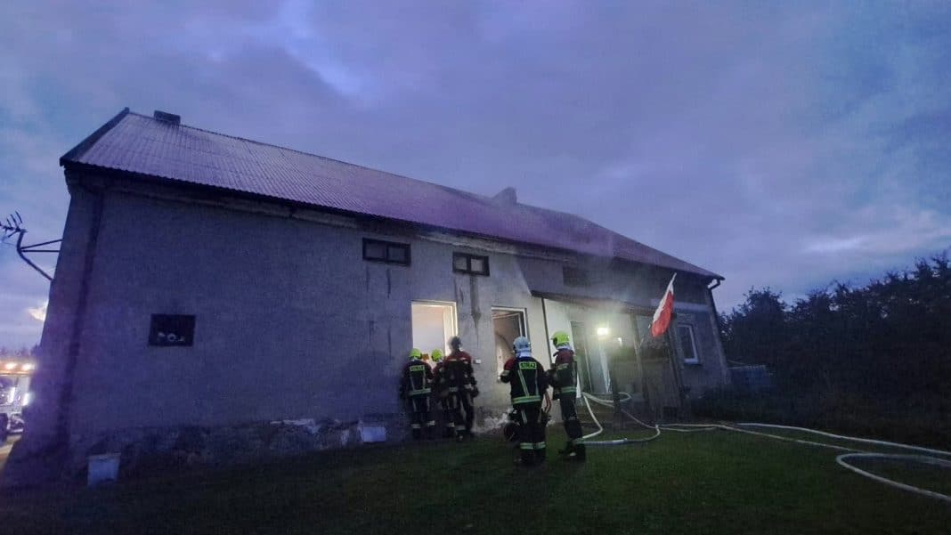 Strażacy gaszą pożar w domu po wybuchu gazu pod Olsztynem, w tle zachmurzony niebo.