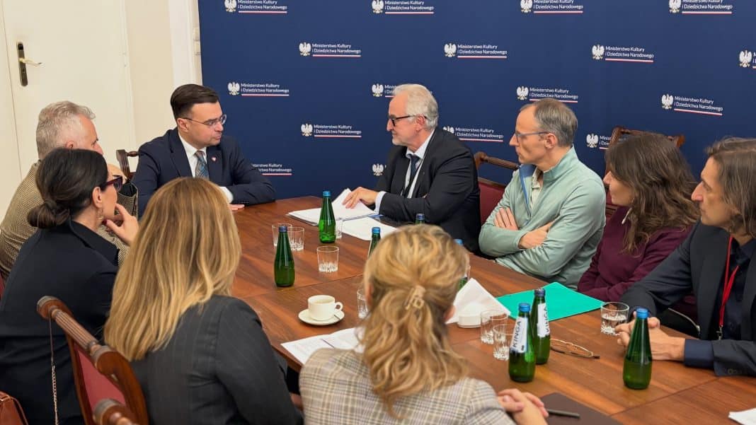 Grupa osób rozmawia przy stole w sali konferencyjnej z logo Ministerstwa Kultury i Dziedzictwa Narodowego w tle.