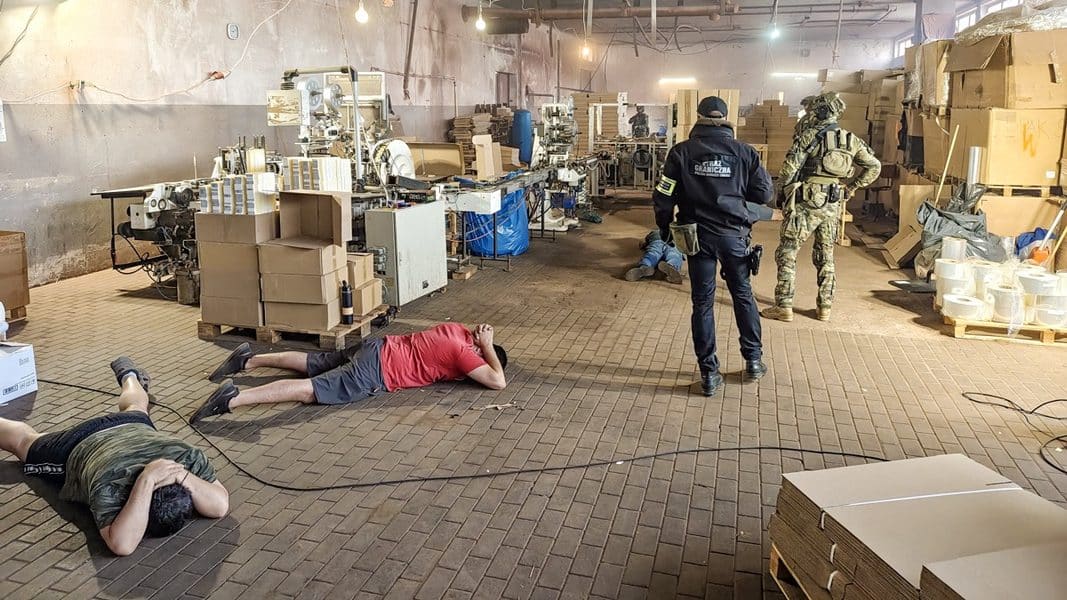Widok na wewnątrz hali z maszynami i kartonami, na podłodze leżą zatrzymani mężczyźni, obecni funkcjonariusze.