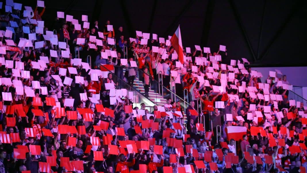 Tłum kibiców w hali Urania, trzymających czerwono-białe flagi i kartki, wspierających reprezentację.