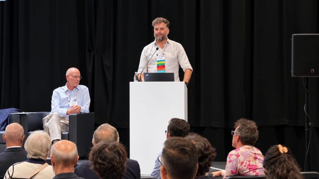Mężczyzna przemawia na konferencji przed zgromadzoną publicznością, za nim czarna kurtyna.