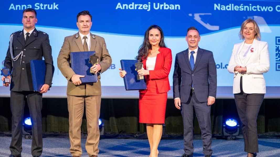 Grupa ludzi na scenie podczas uroczystości, wśród nich strażak z odznaką, trzymający nagrodę i dyplom.