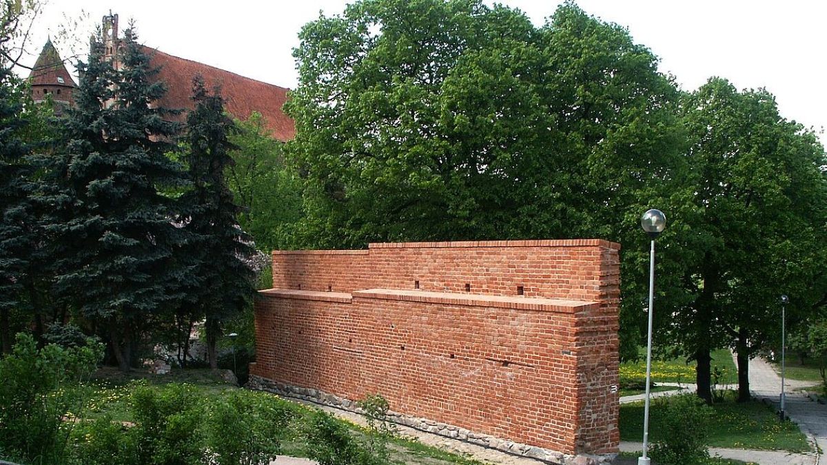 Fragment dawnego muru obronnego w parku z drzewami i latarniami, widoczny na tle zieleni.