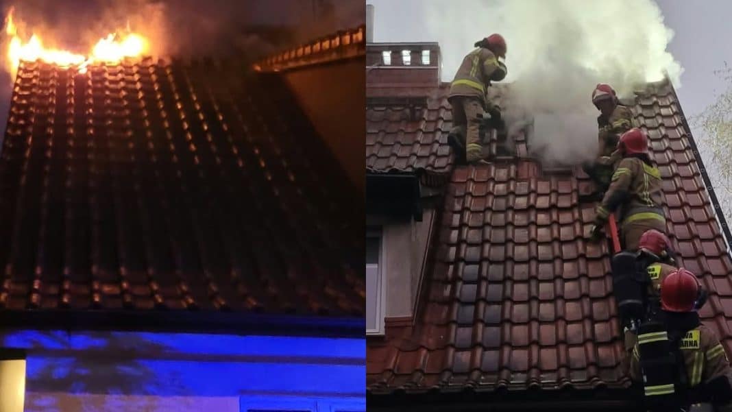 Zdjęcie przedstawia pożar domu w Olsztynie, z widocznym ogniem na dachu oraz strażaków gaszących pożar.